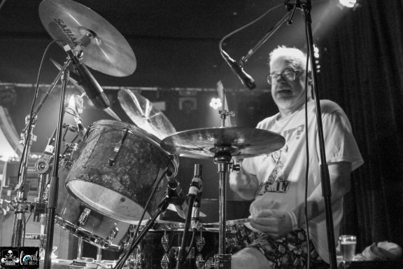 Dead Bob - La Source de la Martinière Black and white photo of a drummer performing on stage, illuminated by stage lights.