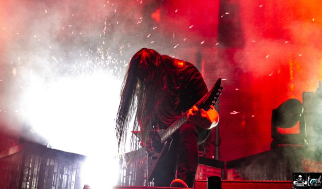 A guitarist with long hair performing in a dramatic, colorful light setting.
