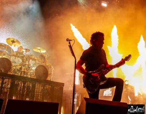 Silhouette of a guitarist on stage with dramatic flames and a drum set in the background.