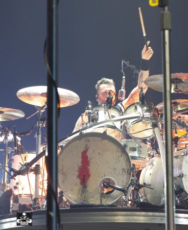 Drummer performing energetically on stage, surrounded by drum set and lights.