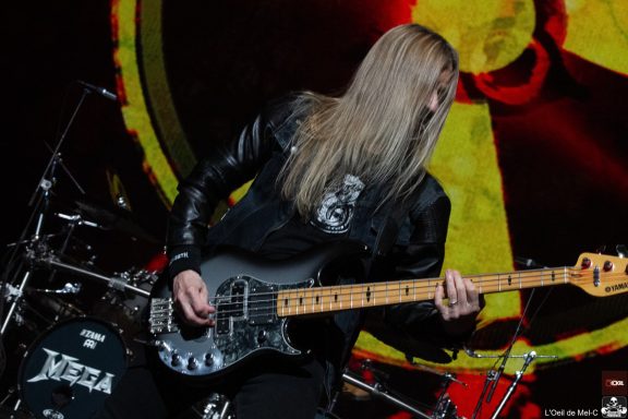 Musician playing a silver bass guitar on stage with a colorful abstract backdrop.