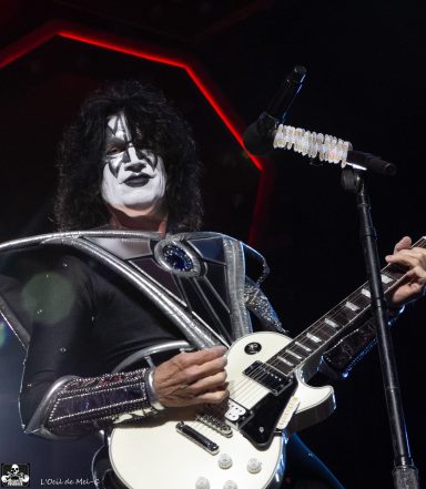 A performer with face paint and a metallic costume plays a white guitar on stage.