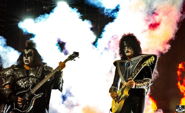 Two musicians in elaborate costumes perform on stage with bright pyrotechnics in the background.