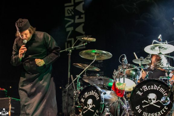 A musician performs on stage with drums and a dramatic backdrop.
