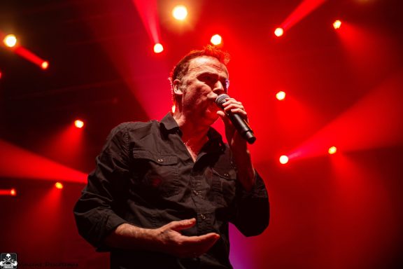 Man singing into a microphone on stage with red spotlight beams in the background.