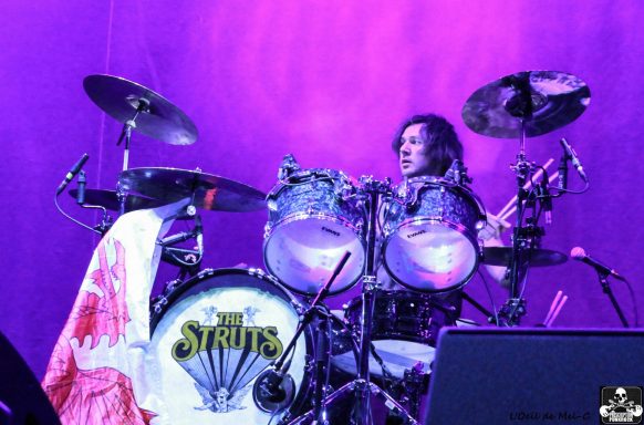 Drummer performing on stage with vibrant purple lighting and a white towel on the drum set.
