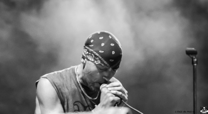 A musician performs on stage, wearing a bandana and singing into a microphone.