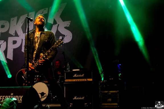 Lead singer performing on stage with guitars and bright green lighting in the background.