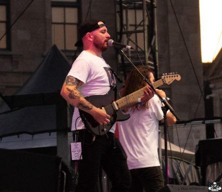 Man playing electric guitar on stage with a singer in the background at a music festival.