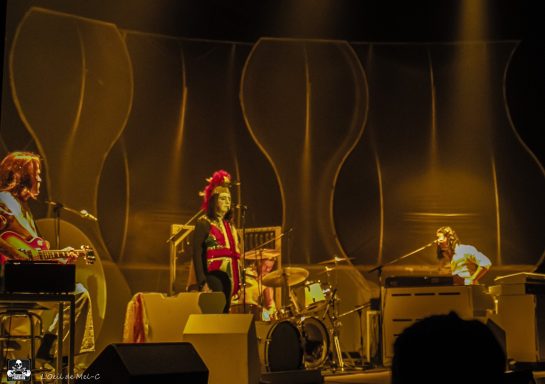 Three musicians performing on stage with colorful lighting and abstract backdrop.