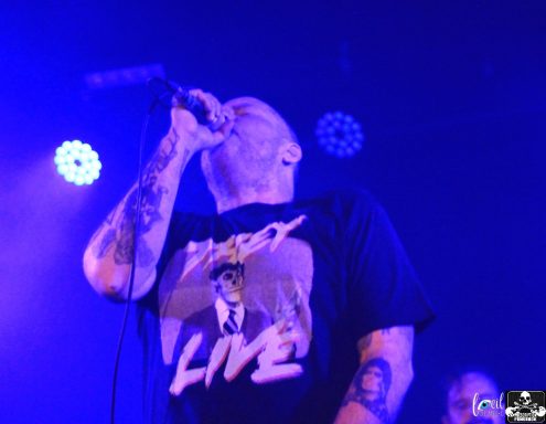 Vocalist performing passionately on stage with purple lighting and visible tattoos.