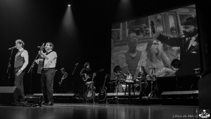 Two singers perform on stage, with a black and white backdrop featuring a scene from a film.