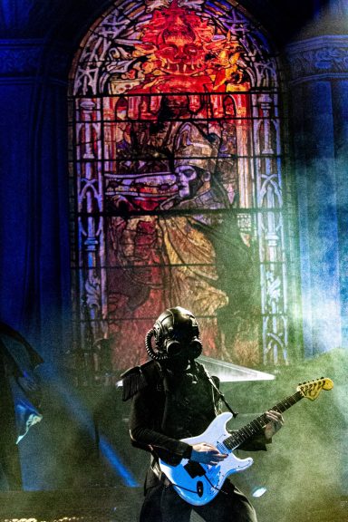 A guitarist in a dark outfit performs in front of a colorful stained glass backdrop.