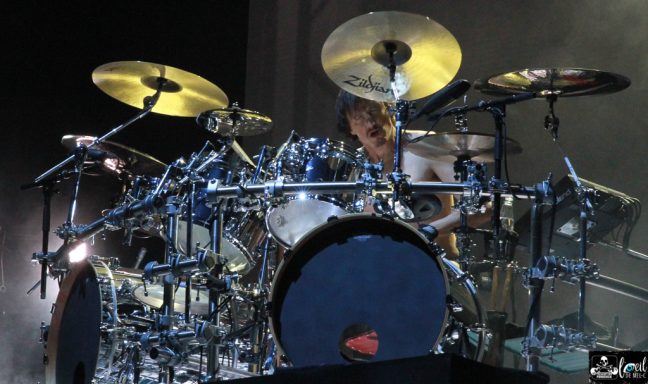 Drummer performing on stage, surrounded by multiple cymbals and a vibrant setup.