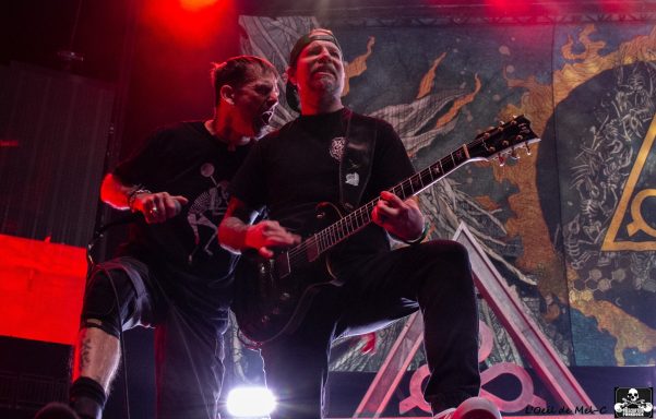 Two guitarists perform energetically on stage with vibrant lights behind them.