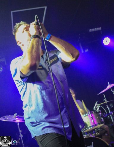 Lead singer performing passionately on stage with purple lighting and a drum set in the background.