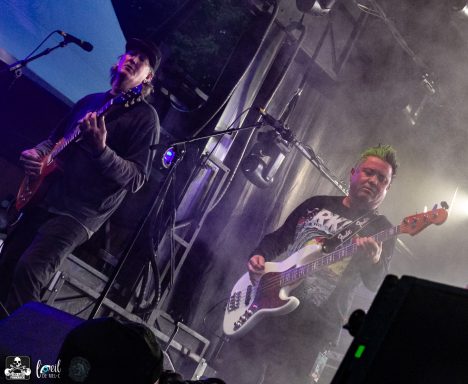 Two musicians performing onstage with electric guitars and smoke effects.