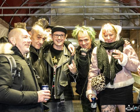 Five people pose together, striking playful poses and smiling, with vibrant clothing and hairstyles.