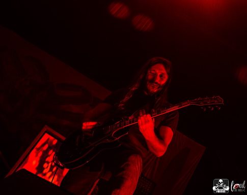 A guitarist performing on stage under red lighting.