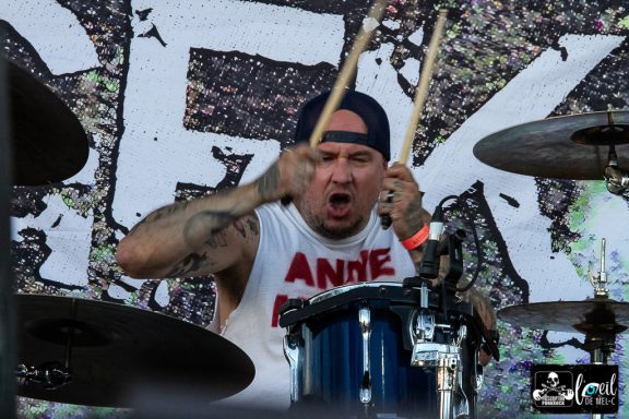 Drummer passionately performing on stage, wearing a tank top and holding drumsticks.