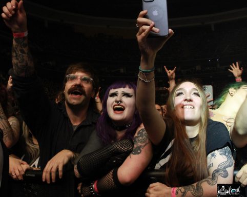 Crowd of enthusiastic concert-goers with various tattoos, cheering and taking pictures.