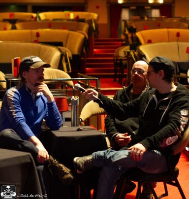 Two men interview a seated man in a theater with empty chairs in the background.