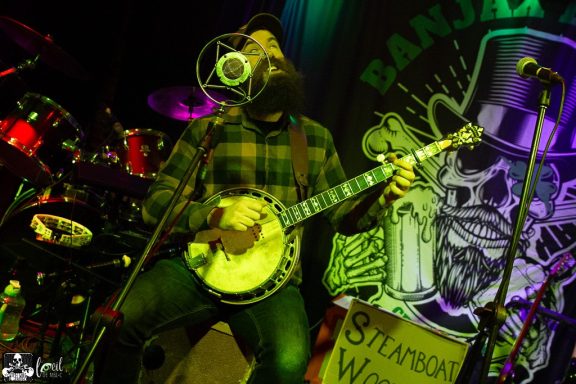 Musician playing a banjo on stage with colorful lights and a backdrop.