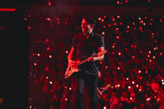 Bryan Adams - Canada Musician playing guitar on stage with a crowd illuminated by red lights.
