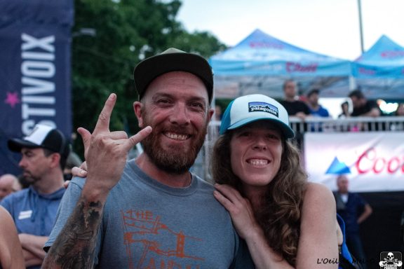 A man making a hand gesture stands beside a woman, both smiling at an outdoor event.