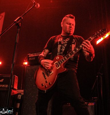 Musician playing electric guitar on stage with red lighting.