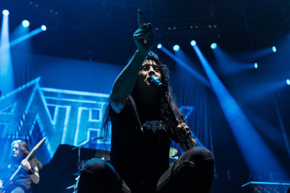 A performer on stage passionately singing with a raised finger, spotlighted in blue light.