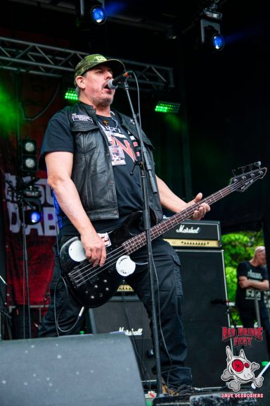 A musician in a black vest plays electric bass on stage at a concert.