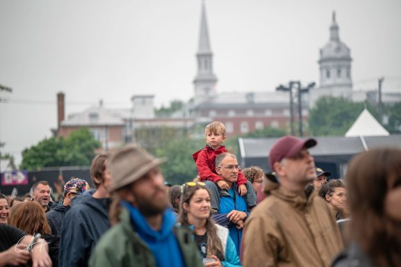 Crowd at an outdoor event with a child on a parent's shoulders and buildings in the background.
