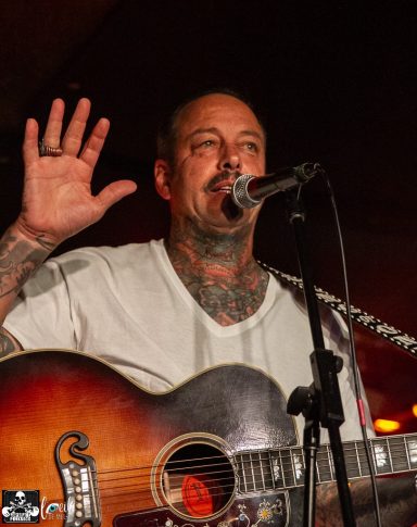 Musician with tattoos sings and plays guitar, raising one hand while addressing the audience.