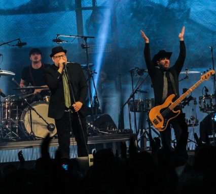 A band performs on stage with a drummer, singer, and bassist, engaging the audience.