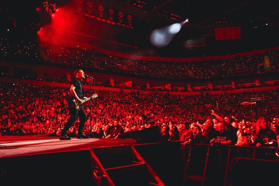 A musician performs on stage in front of a large, cheering crowd with red lighting.