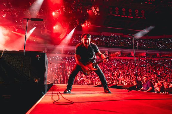 A guitarist performing energetically on stage with red stage lights and a large crowd.