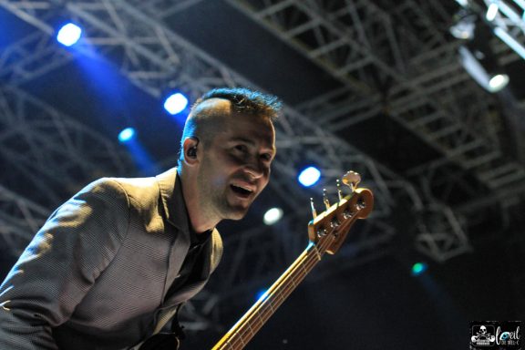 A musician playing bass guitar on stage, smiling under stage lights.