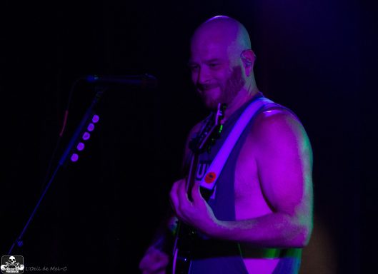 A muscular bald man playing guitar on stage with colorful lighting behind him.
