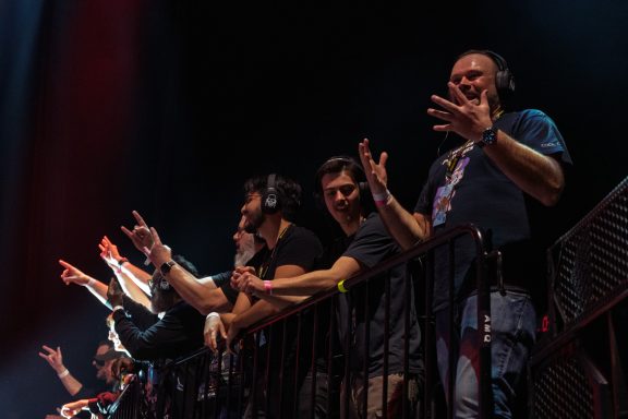 Crowd cheering and gesturing from a concert balcony, wearing headphones.