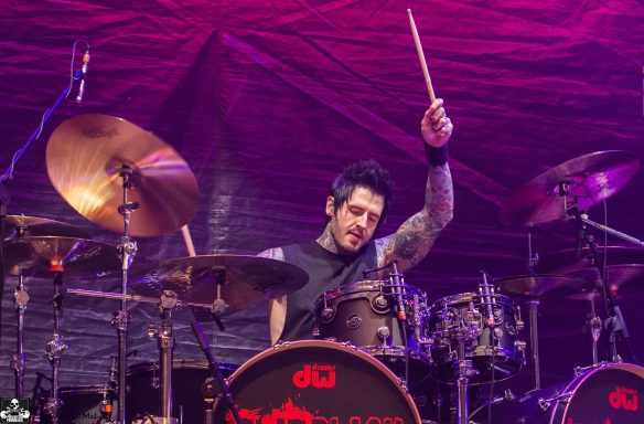 Drummer energetically performing on stage with colorful lighting behind.