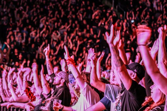 Crowd at a concert with raised hands, illuminated by colorful stage lights.