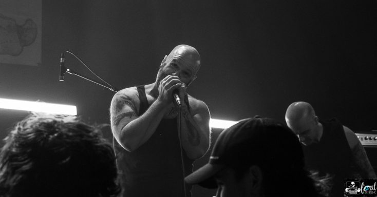 Male singer performing on stage, holding a microphone, with a blurred audience in shadow.
