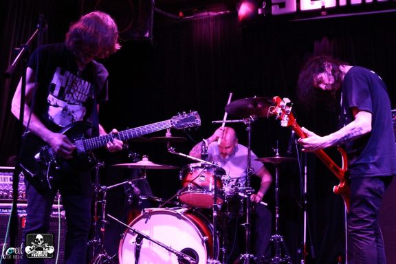 Band performing on stage with guitarists and a drummer in a dimly lit venue.