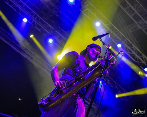 Musician playing saxophone on stage with vibrant blue and yellow lights.