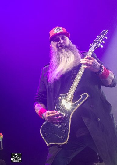 A musician with a long beard and red cap plays electric guitar on stage.