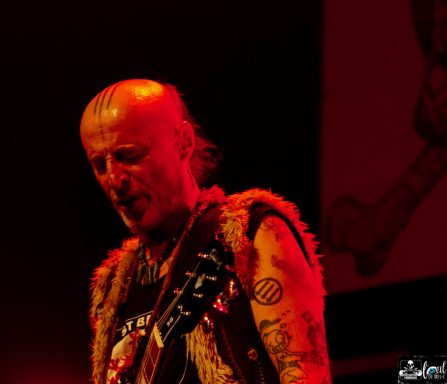 Musician playing guitar on stage, wearing a fur vest, with red lighting and tattoos visible.