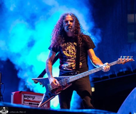 Male guitarist on stage with long curly hair, holding an electric guitar amid blue lighting.