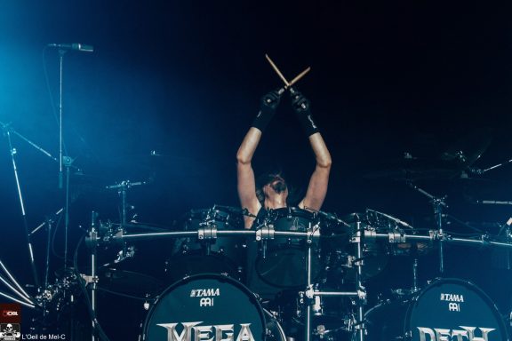 Drummer performing energetically on stage, raising drumsticks above their head.