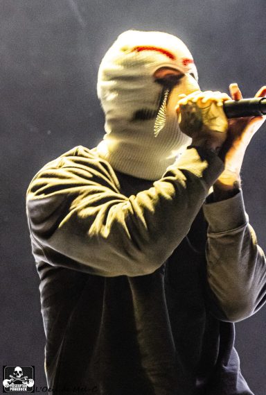 Performing artist in a white mask, holding a microphone on stage.
